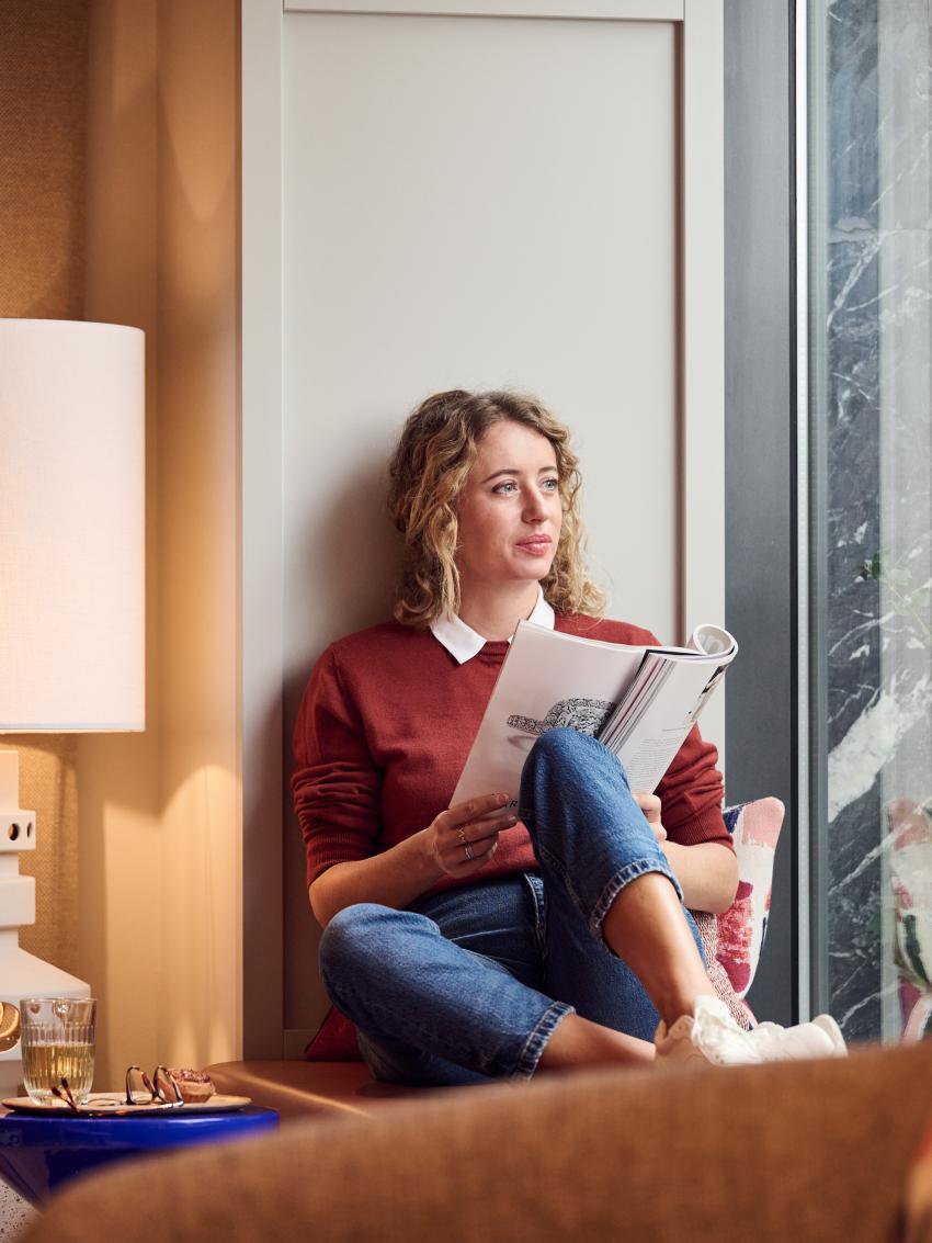 Frau liest ein Magazin auf einem Fenstersims und blickt durch ein Fenster
