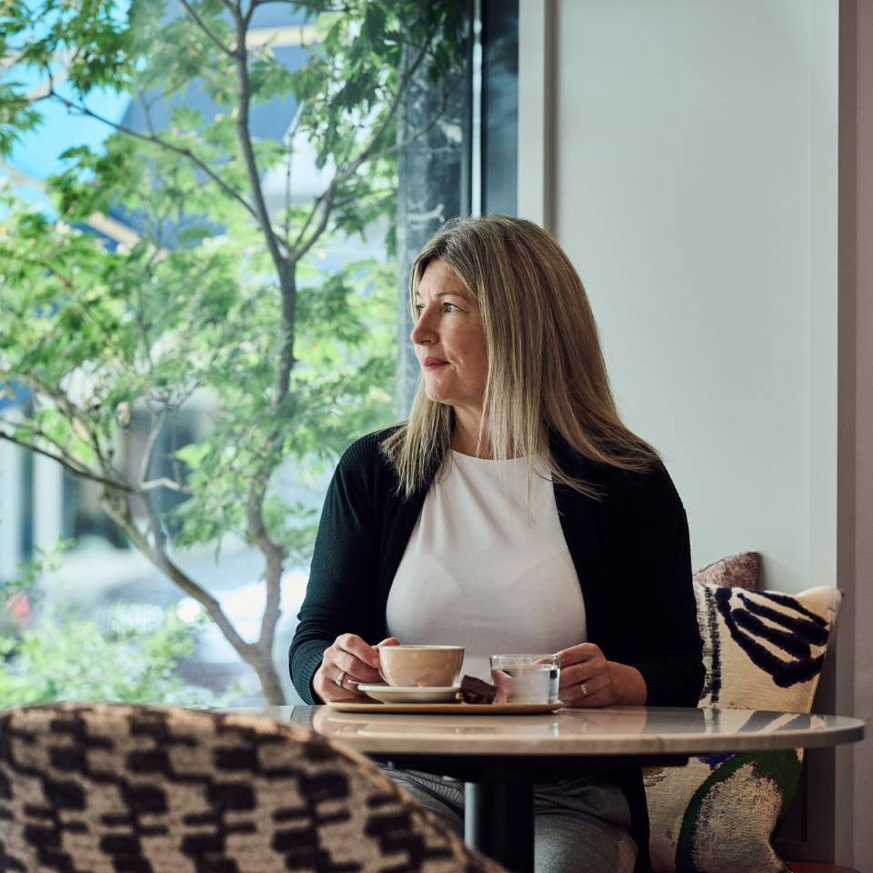 Frau sitzt an einem Tisch beim Fenster und trinkt Kaffee
